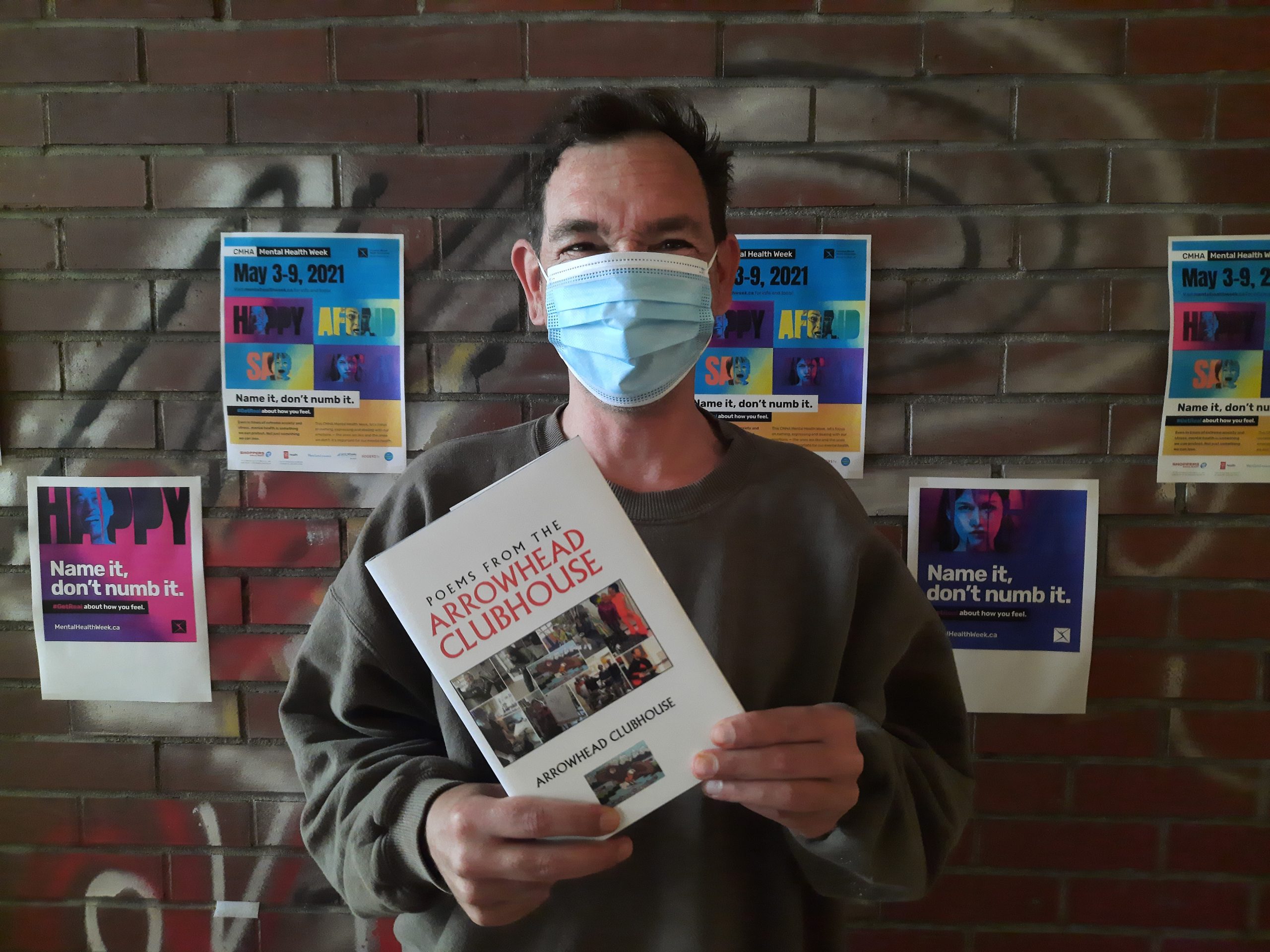 A man in a mask holds a book that says "Poems from the Arrowhead Clubhouse", against a brick wall with posters on it.